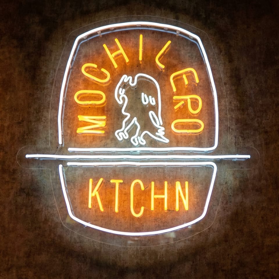 Neon sign reading “Mochi Frog Kitchen,” featuring a stylized frog on a dark textured wall.