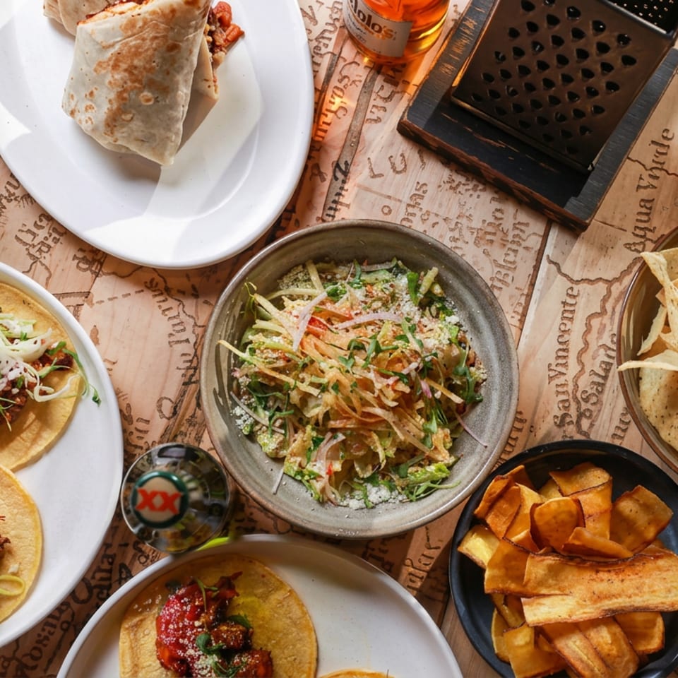 Overhead spread of Mexican food including tacos, burritos, salsa, tortilla chips, rice, and salad on a rustic table.