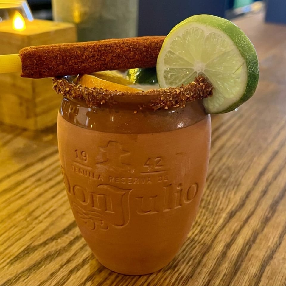 Cocktail in a clay cantarito glass with chili-salt rim, lime slice, orange wedge, and cinnamon stick garnish on a wooden bar.