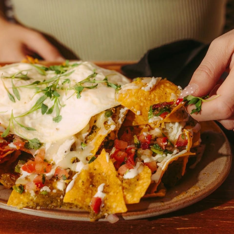 Loaded nachos topped with melted cheese, salsa, herbs, and sour cream, while a diner lifts a chip beside a rimmed cocktail.