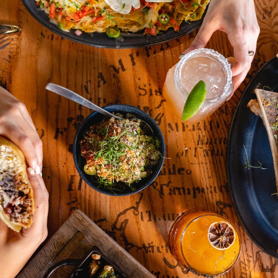 Overhead restaurant table spread featuring tacos, nachos, flatbread, dipping sauces, colorful cocktails, and hands sharing food.