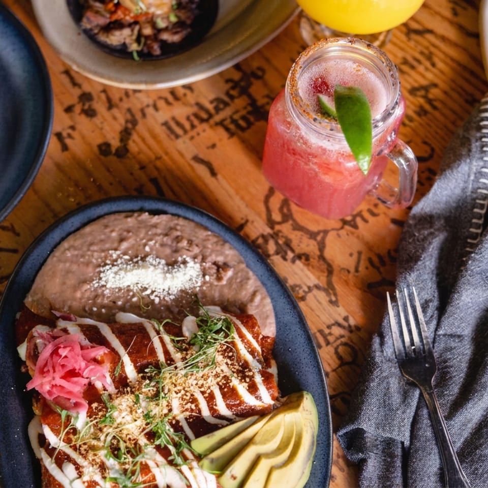 Overhead view of Mexican dishes, including enchiladas, beans, soup, garnishes, and a pink lime cocktail on a wooden table.