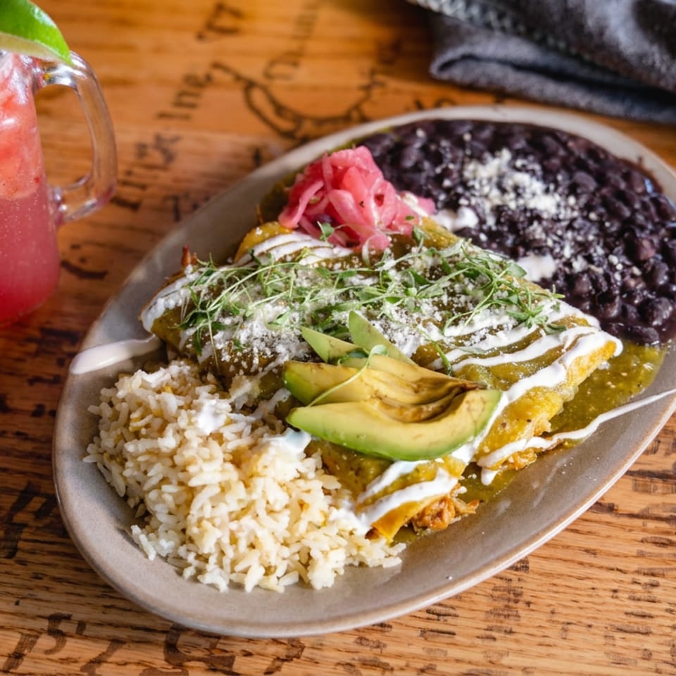 Mexican-inspired enchilada plate with rice, black beans, avocado, pickled onions, and crema beside a pink lime drink.