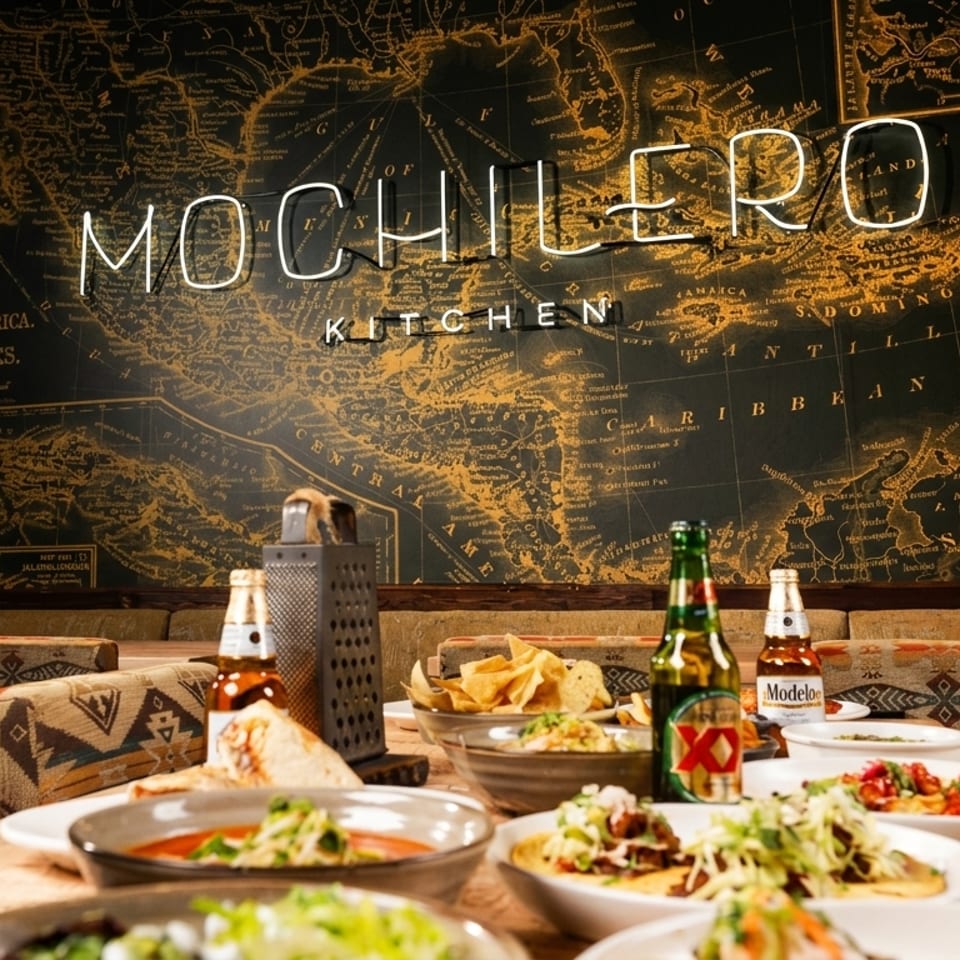 Restaurant table displays Mexican dishes, chips, and beer beneath a large vintage map mural with Mochilero Kitchen signage.