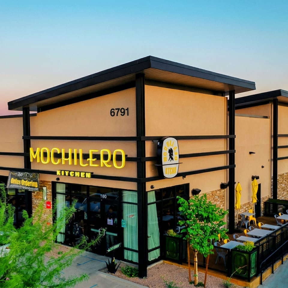 Mochilero Kitchen restaurant exterior with yellow signage, covered patio seating, umbrellas, and landscaped surroundings.