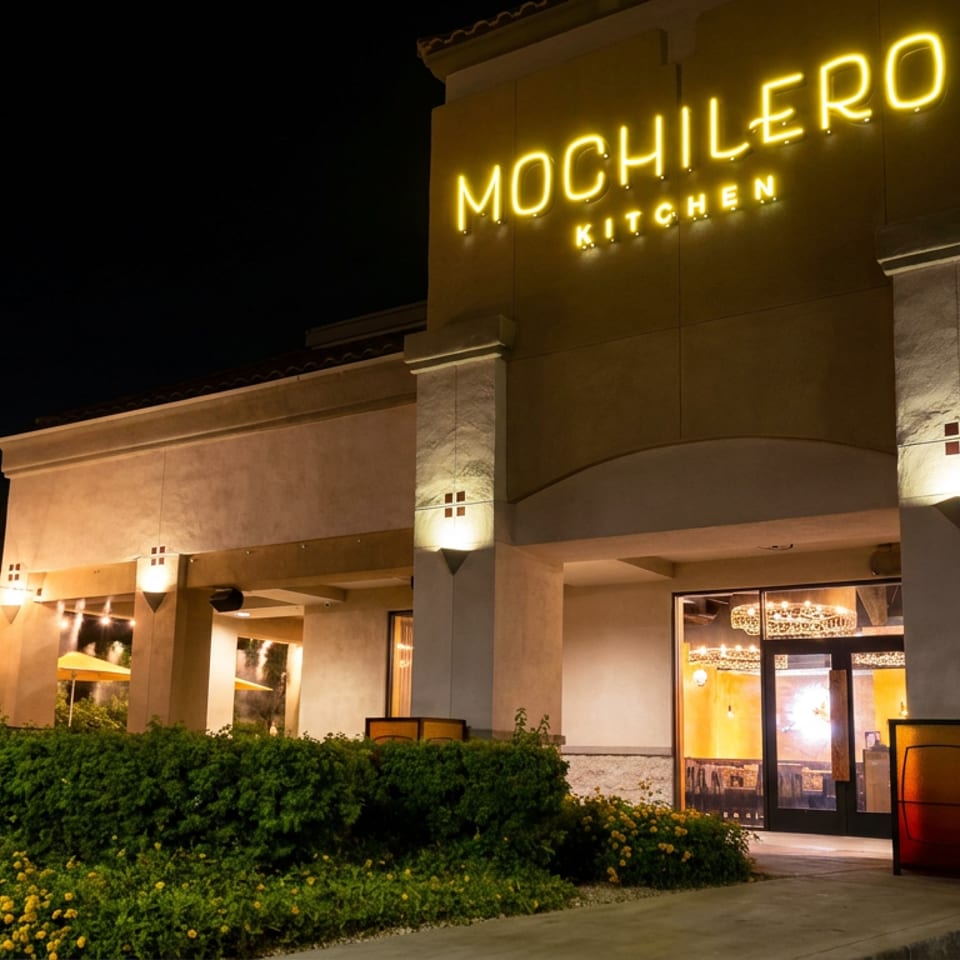 Mochilero Kitchen restaurant exterior at night, with glowing neon signage, illuminated patio umbrellas, landscaping, and warmly lit entrance.