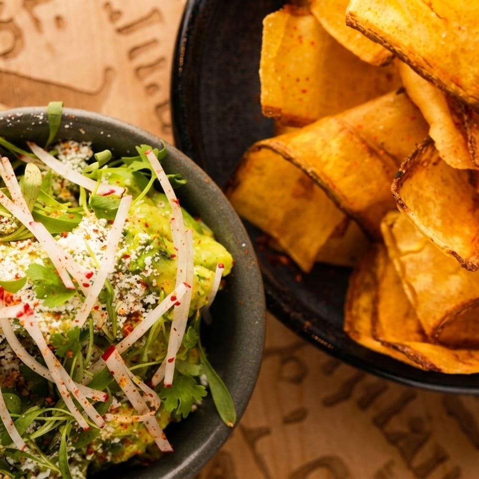 Guacamole topped with herbs, crumbled cheese, and radish beside a bowl of golden tortilla chips on a wooden table.